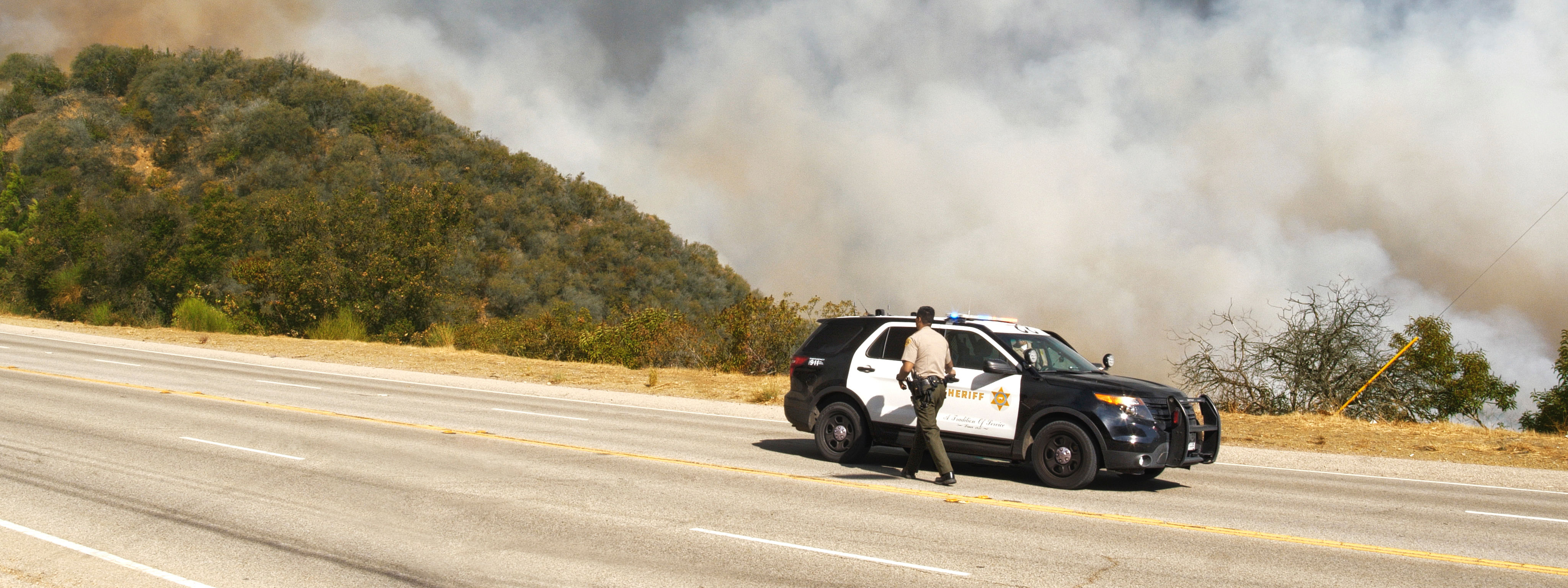 A sheriff's deputy walking near a patrol vehicle on a highway, with a large wildfire and thick smoke covering the hillside in the background.